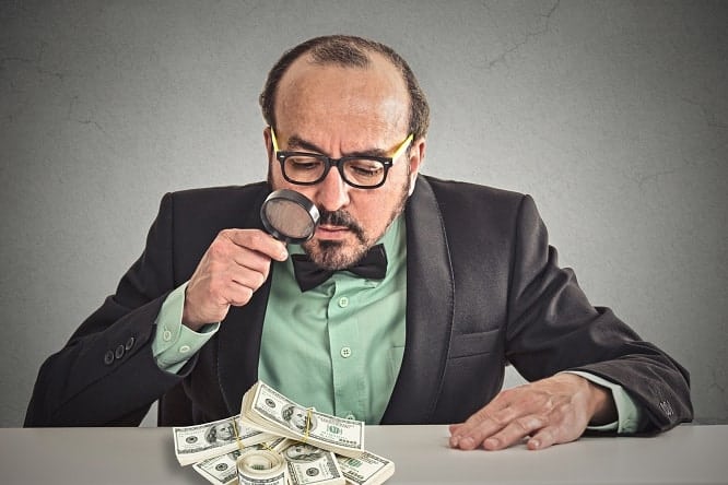 penny pincher. Business man looking through magnifying glass on stack of dollar banknotes on table isolated grey office background. Economy financial wealth success concept. Ponzi scheme investigation